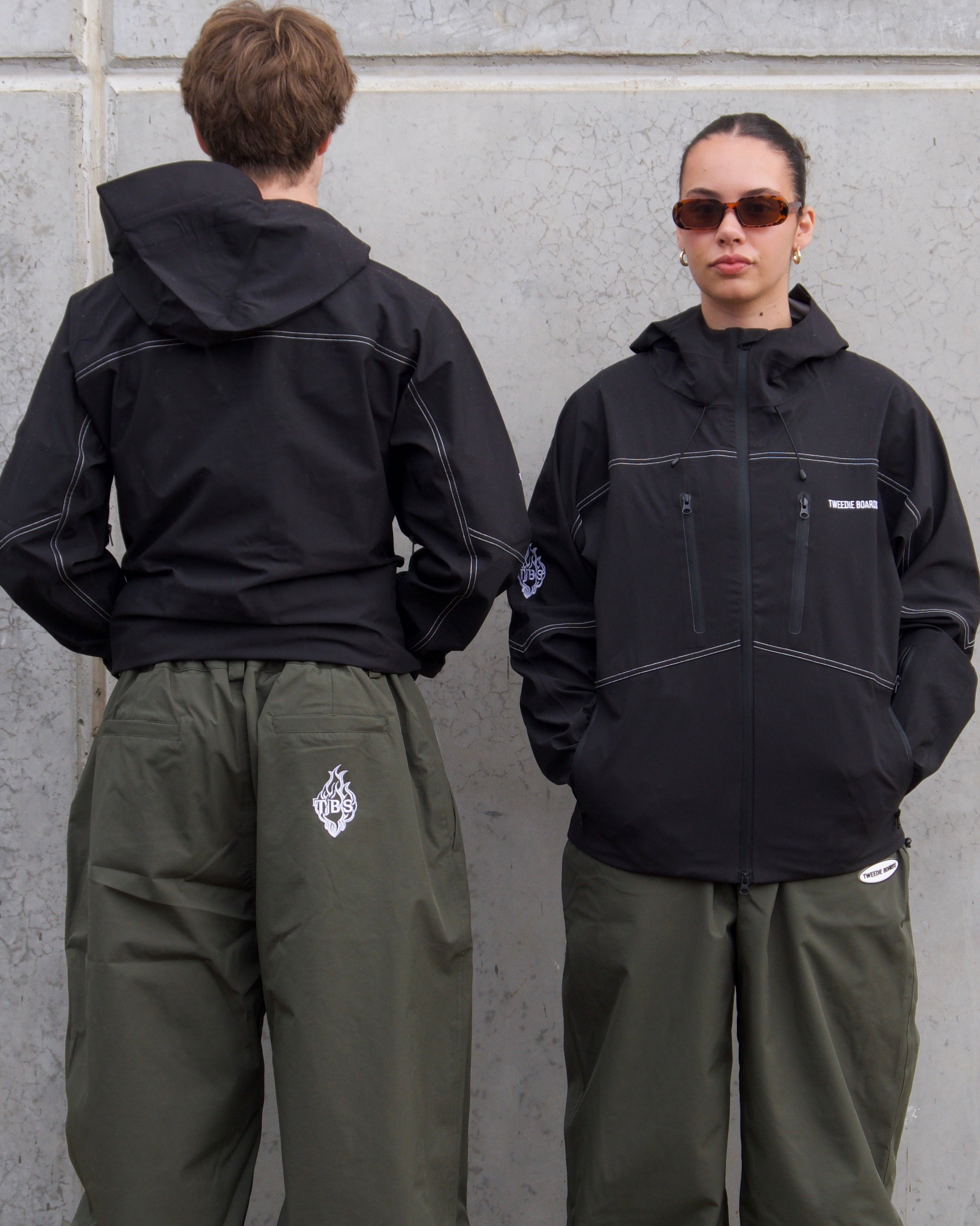 Two people wearing black jackets and green pants standing against a gray wall.