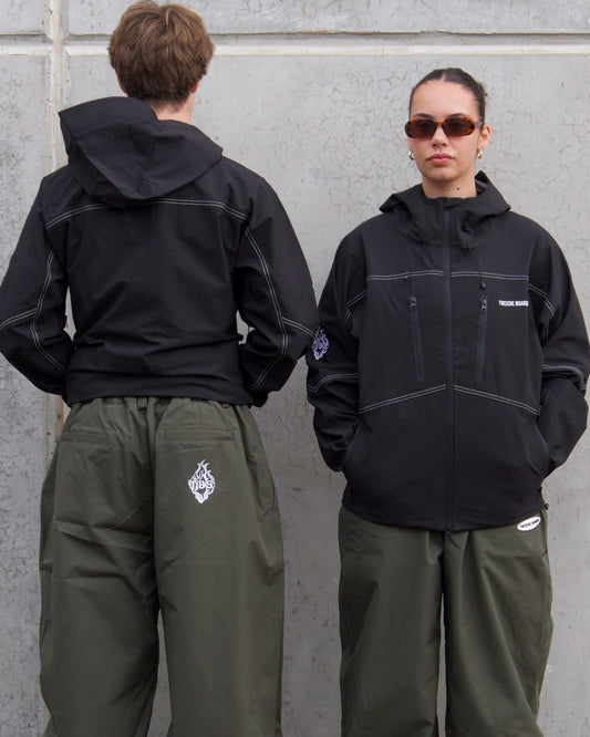 Two people wearing black jackets and green pants standing against a gray wall.