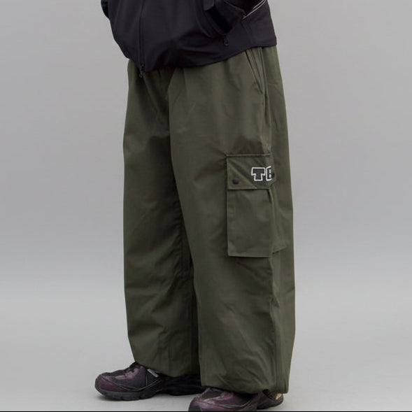 Person wearing a black jacket and green cargo pants standing against a gray wall.