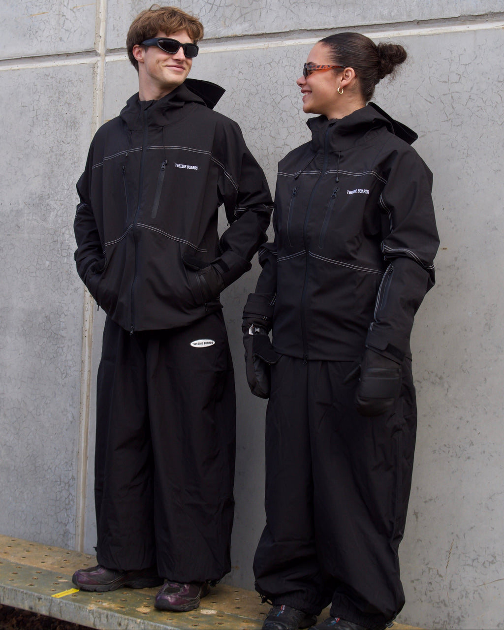 Two people wearing black outfits standing against a gray wall.