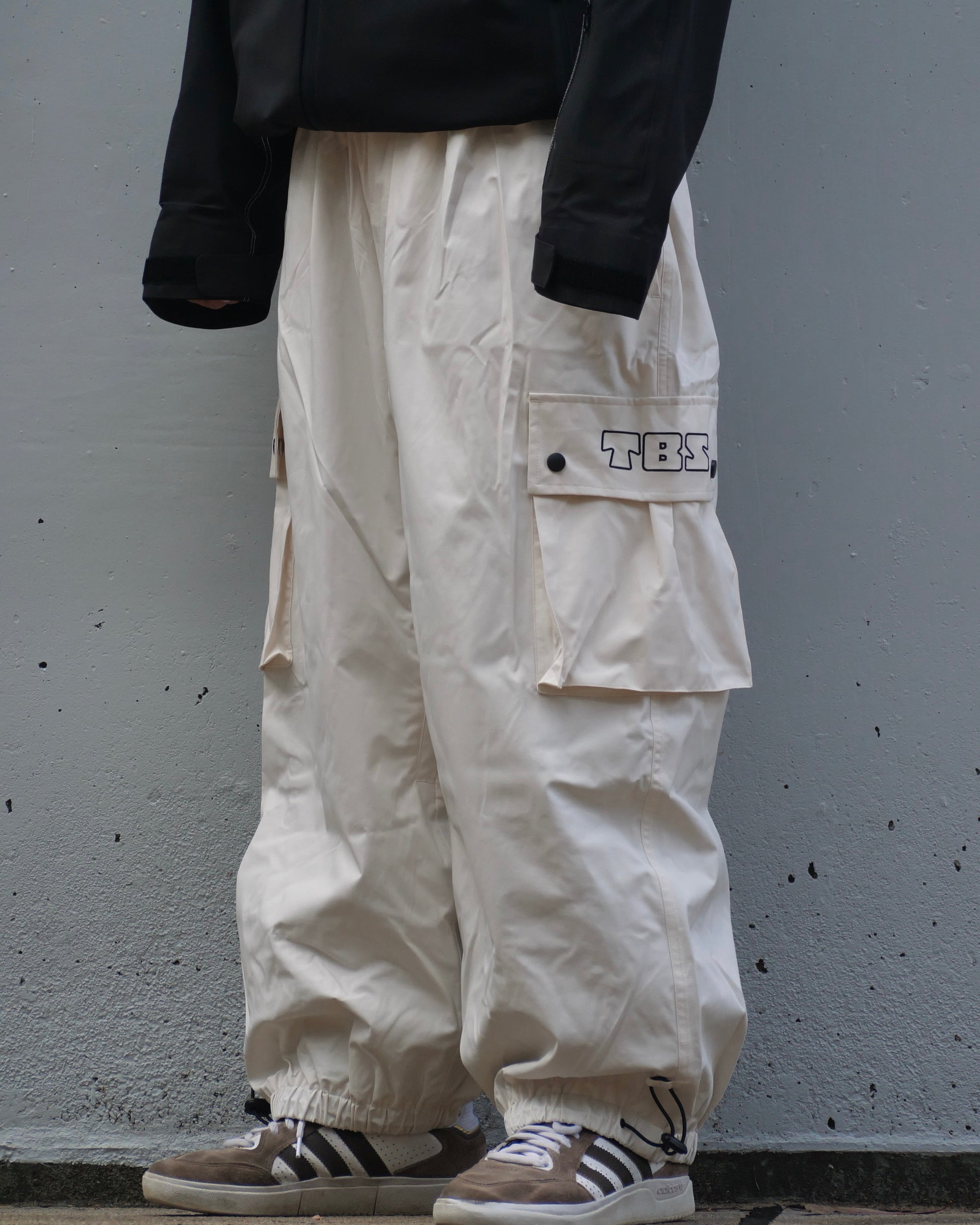 Person wearing white cargo pants with a black jacket and Adidas shoes against a gray wall.