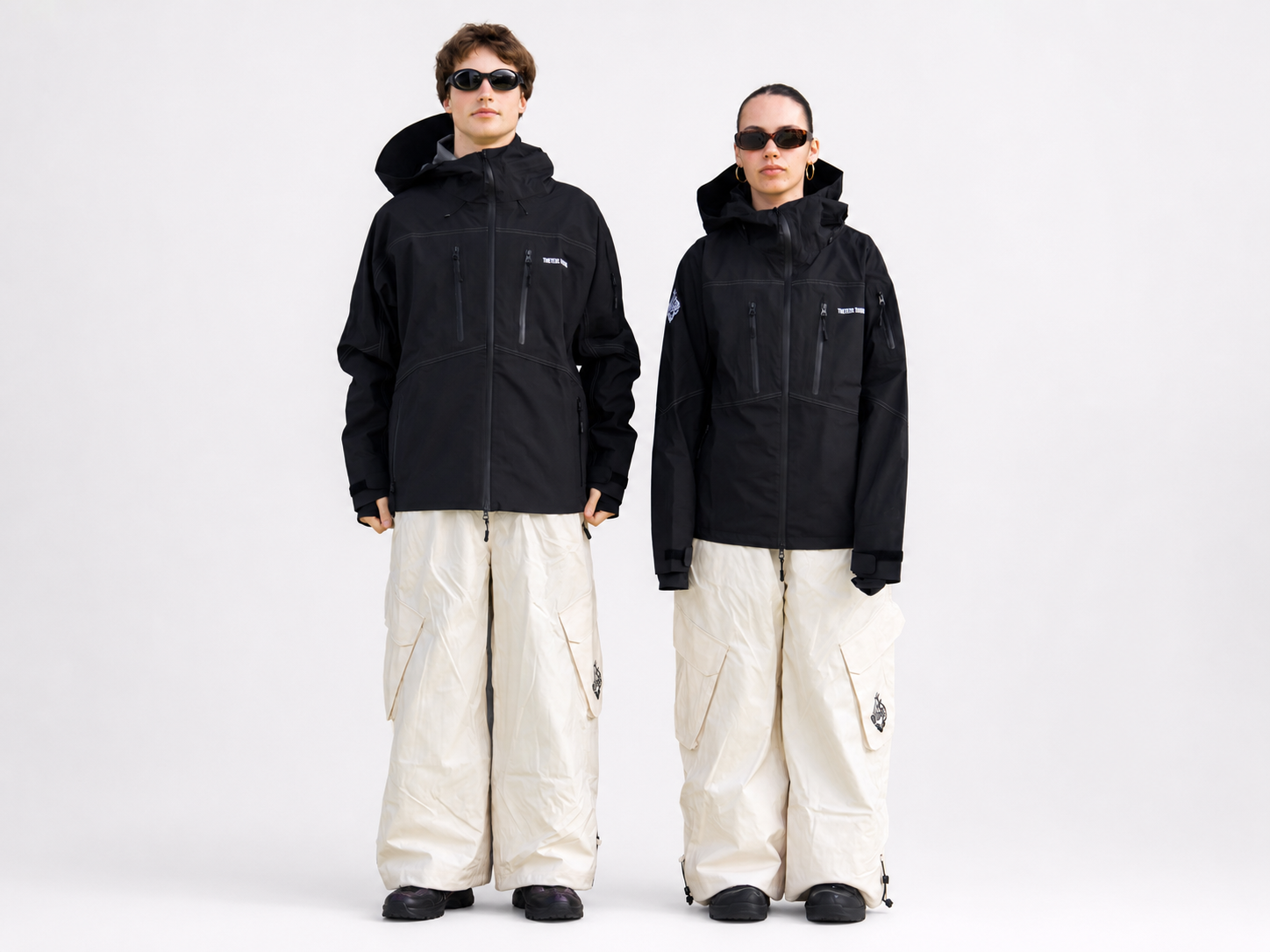 Two models standing side by side wearing black hooded technical jackets and off-white wide-leg cargo pants against a plain light gray studio background.