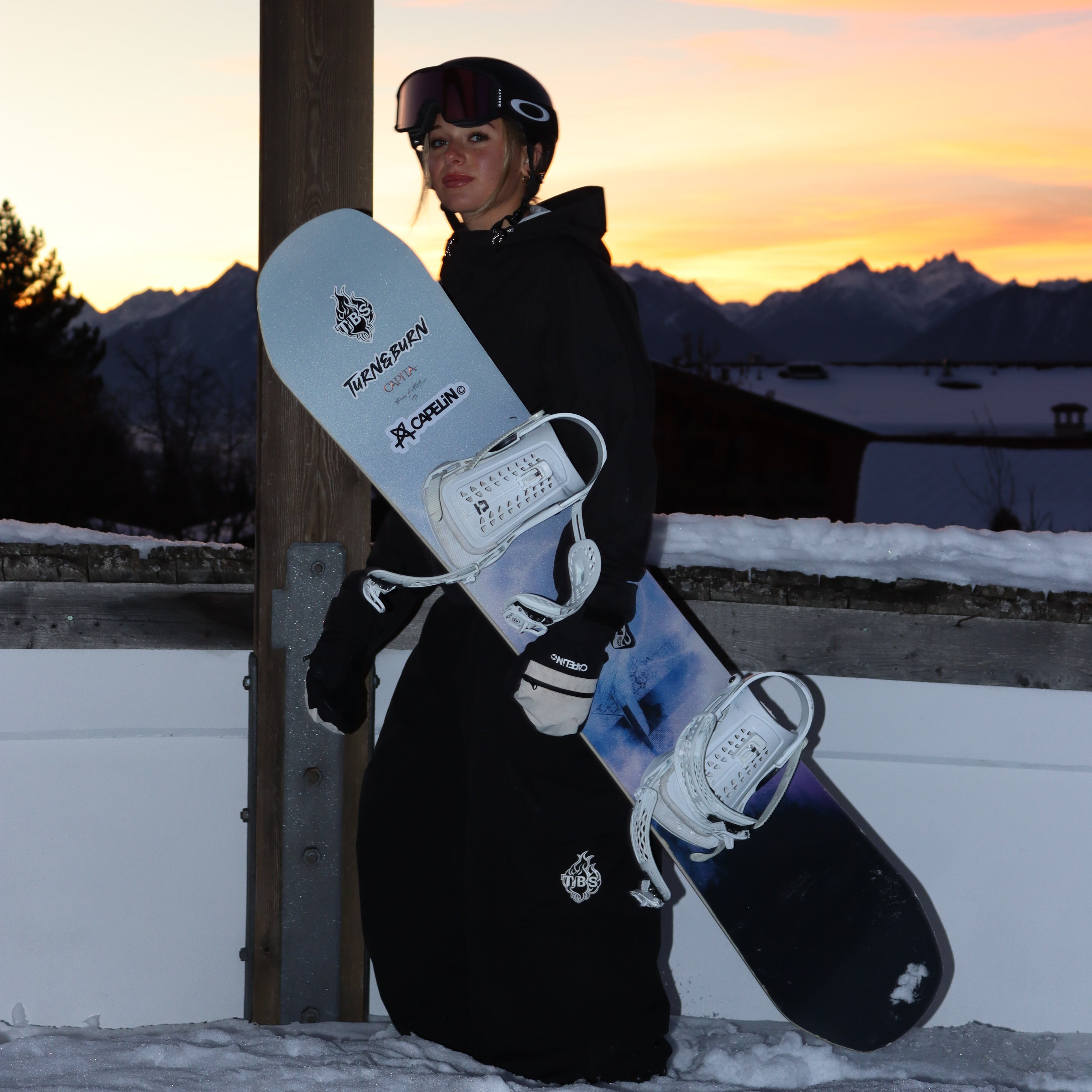 Person holding a snowboard against a sunset sky with mountains in the background