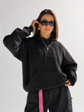 A woman modeling a black oversized hoodie with a tonal black logo on the chest, styled with black sweatpants featuring a pink side stripe, black sunglasses, and a silver chain necklace against a light gray background.