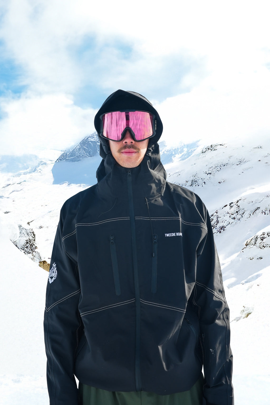 A person standing in a snowy mountain landscape wearing a black hooded ski jacket, dark pants, and pink-tinted ski goggles. The jacket has subtle light piping details and a small white logo on the chest.