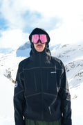 A person standing in a snowy mountain landscape wearing a black hooded ski jacket, dark pants, and pink-tinted ski goggles. The jacket has subtle light piping details and a small white logo on the chest.