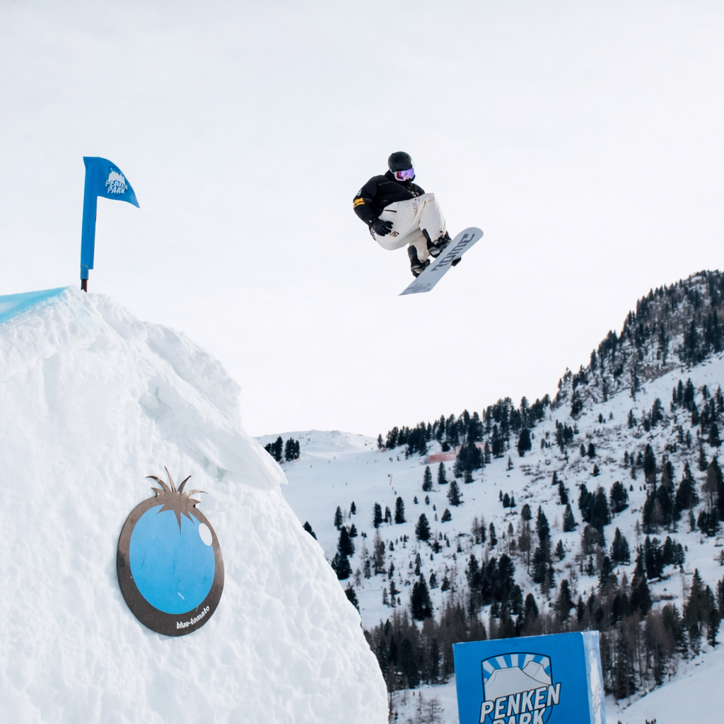 Snowboarder performing a trick in the air with a snowy mountain and Penken Park logo in the background.