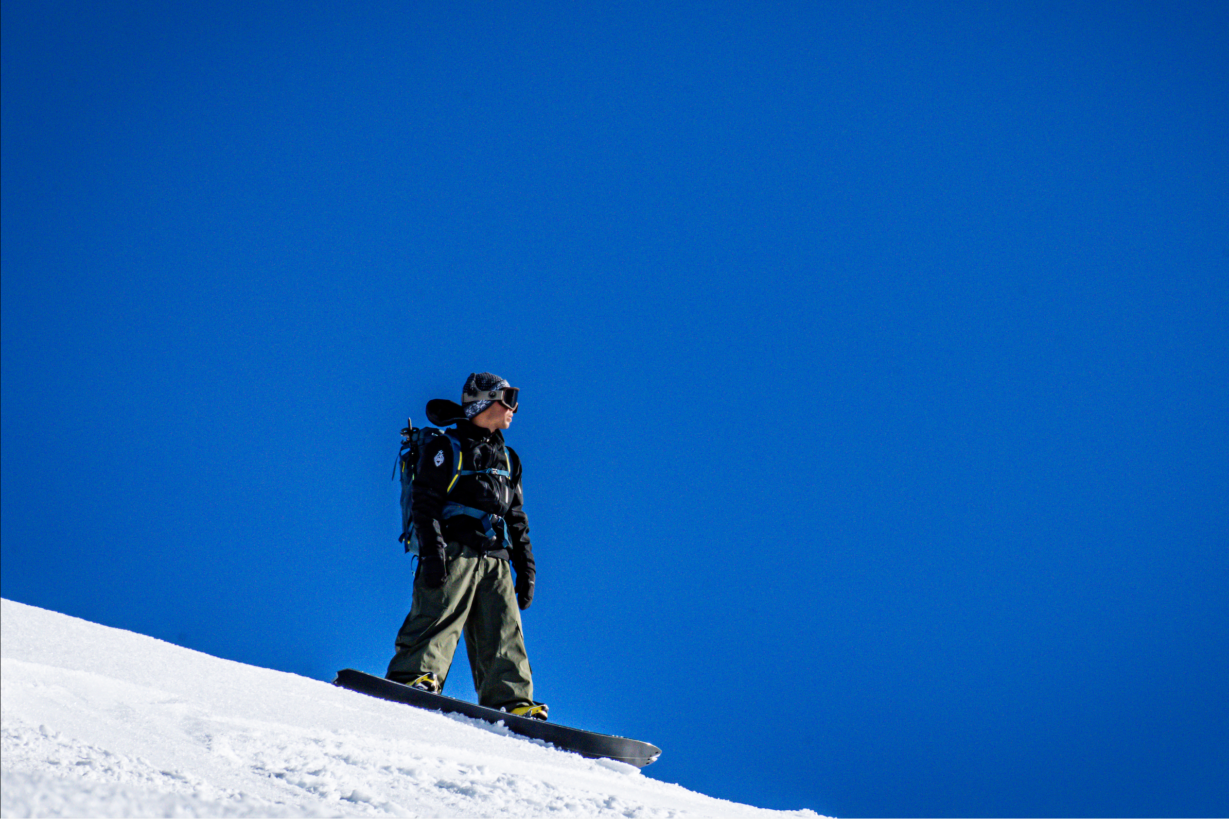 A snowboarder stands on a snowy slope under a vivid blue sky, wearing dark winter gear and goggles.